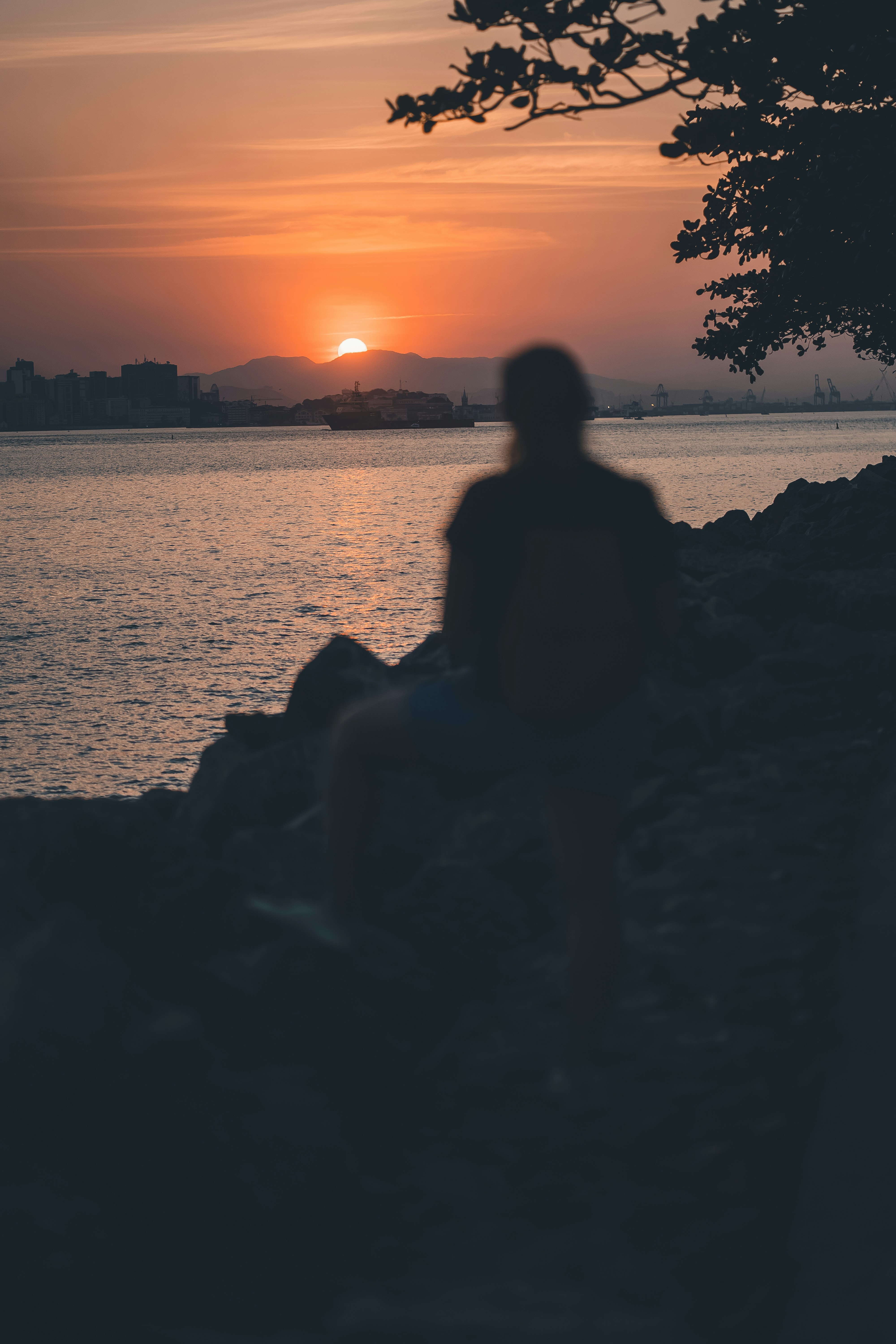 silhouette of woman sitting on rock near body of water during sunset
